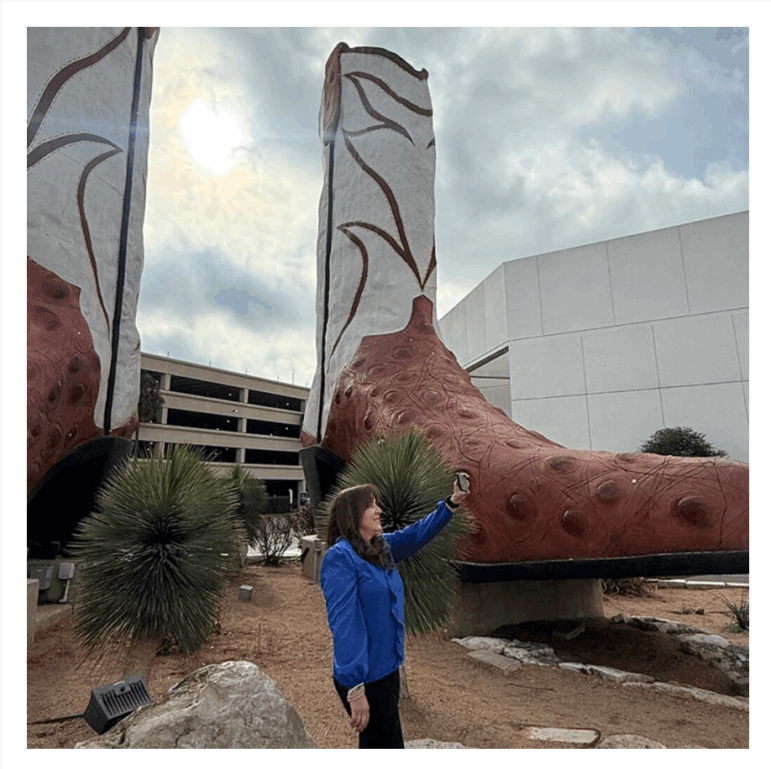 A person stands in front of a statue of an enormous pair of cowboy boots and appears to be taking a selfie.